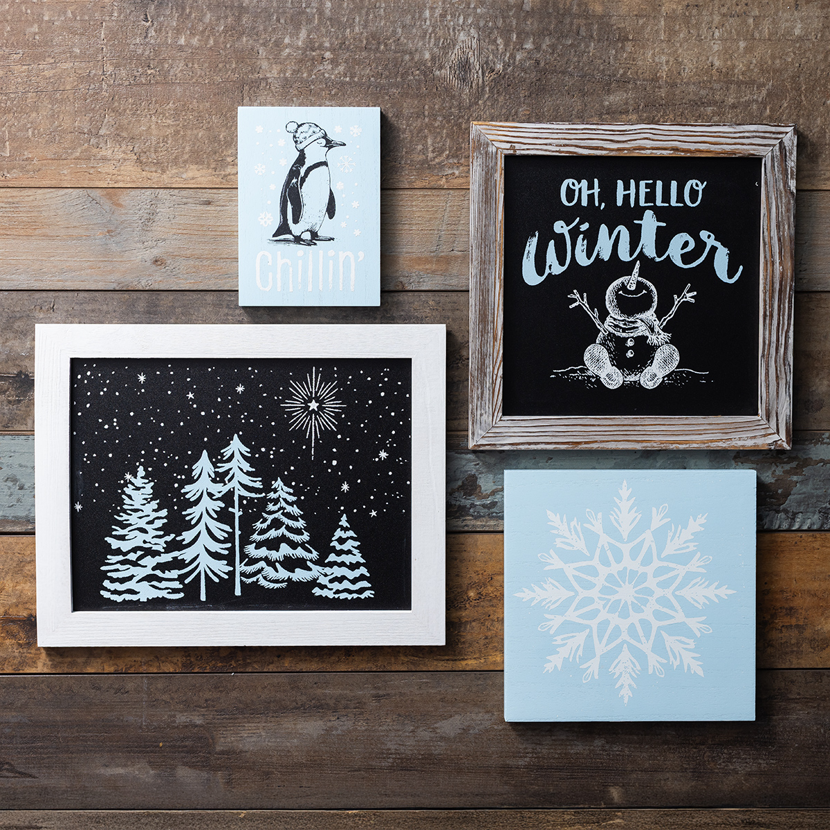 Four winter-themed wall art pieces on a wooden wall: a penguin with "chillin", "Oh, Hello Winter" with a snowman, a snowy forest, and a blue snowflake on a pale blue background.