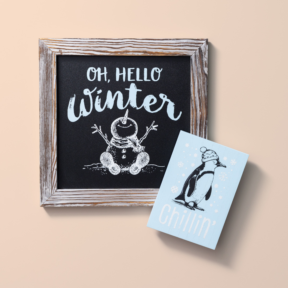 A framed chalkboard with "Oh, Hello Winter" and a snowman drawing sits next to a blue card featuring a penguin, snowflakes, and the word "Chillin'" on a beige background.