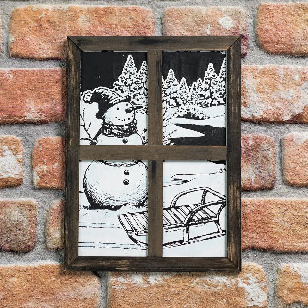 A framed black and white drawing of a snowman, snowy trees, and a sled, designed to look like a window, is mounted on a brick wall.
