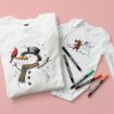 Two white garments, a T-shirt with a snowman and cardinal illustration, and a baby onesie with a mouse building a snowman, are laid on a pink background. Four colored markers are placed next to them.