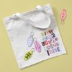 A white tote bag with colorful, cut-out style text reading "Slow down, you’re doing fine," surrounded by four motel keychains with various phrases, on a light yellow background.