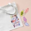 A white tote bag with pastel text reading “Slow down you’re doing fine” lies next to four colorful motel-style keychains with playful phrases, all on a pale pink background.