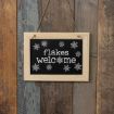 A small blackboard sign with a light wood frame hangs on a rustic wooden wall. The sign says “flakes welcome” in white letters, with snowflakes surrounding the text and a large snowflake replacing the "o" in "welcome.