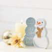 A decorative snowman figure, a gold ornament, a cream-colored flower, and a lit candle are arranged on a white surface with a sign that reads "Hello Winter, Let it snow, Merry Christmas.