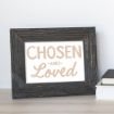 A dark wooden picture frame with a white print inside that reads "CHOSEN AND Loved" in beige and light brown letters, displayed on a light wooden surface next to a small stack of books.