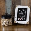 A glass jar labeled "Oh Biscuits!" filled with dog biscuits sits next to a framed sign that reads, "This home is filled with kisses, wagging tails, wet noses and love," on a wooden surface.