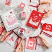 A flat lay of Valentine’s Day cards, small candy boxes, gift tags with hearts and cupids, and a decorative garland with red and pink ribbons and beads, all arranged on a white surface.