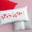 Three pillows on a pink surface: a red pillow, a gray pillow with a white stripe, and a white pillow featuring a design of red and pink heart-shaped balloons.