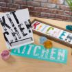 A display of three items featuring the word "KITCHEN" in large, distressed letters: a white sign, a teal mat, and a colorful framed sign, with open paint containers on a wooden table.