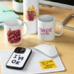 A desk with mugs, a bottle, a phone, and sticky notes. Mugs read “Women always yes oops,” “nope,” and “Older, wiser, hotter than ever.” The phone case says “I’m OK,” and a note reads “That sounds like a tomorrow problem.”.