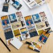 A collection of vintage matchbox-style printed cards and luggage tags are spread on a wooden surface, along with blank ID cards and a white cloth. The designs feature retro illustrations and decorative patterns.