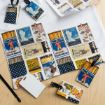A collection of vintage matchbox-style printed cards and luggage tags are spread on a wooden surface, along with blank ID cards and a white cloth. The designs feature retro illustrations and decorative patterns.