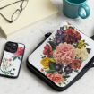 A tablet case with a colorful floral design sits on a marble surface beside a smartphone with a botanical case, a pair of glasses, a closed book, and a turquoise mug.