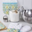 A kitchen scene with a jar of flour and a gold scoop, a stainless steel mixing bowl with a whisk, round cookie cutters, and patterned oven mitts and pot holder on a white surface.