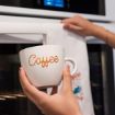 A person holds a white coffee mug with the word "Coffee" written in cursive, while another hand hangs a white towel on an oven handle in a modern kitchen.