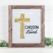 Framed sign with a yellow wooden cross on the left and the words "CHOSEN AND LOVED" in black text on the right, set against a white background with greenery at the bottom.
