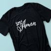 A black t-shirt with the phrase "Can I get an Amen" written in white cursive and bold lettering on the front, displayed on a light blue background.