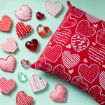 A red pillow with white and pink heart patterns is surrounded by heart-shaped objects, including fabric hearts, glass hearts, and small heart-shaped boxes, all on a light green background.