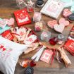 A flat lay of Valentine’s Day crafts on a wooden table, including heart-shaped cards, a garland, a pillow with balloon hearts, small gift boxes with candy, paint jars, and decorative paper cutouts in red, white, and pink.