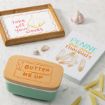 A butter dish, a garlic-themed sign reading "take off your cloves," a penne pasta sign saying "penne for your thoughts," garlic cloves, and dry penne pasta pieces are displayed on a marble surface.