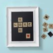 A framed crossword art piece spells "LOVE YOU" with Scrabble-style tiles on a black background. Extra tiles and hearts are scattered nearby on a light blue surface.
