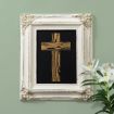 A framed artwork featuring a stylized gold wooden cross on a black background hangs on a green wall. White lilies are placed in the lower right corner of the image.