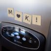 Four wooden Scrabble-style tiles on a stainless steel refrigerator spell "M ♥ K !" above the ice and water dispenser. The tiles include a heart symbol and Scrabble-style numbers.