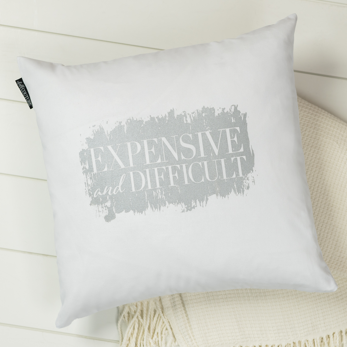 A white square pillow with grey text that reads "EXPENSIVE and DIFFICULT," resting on a cream-colored textured blanket against a white wooden surface.