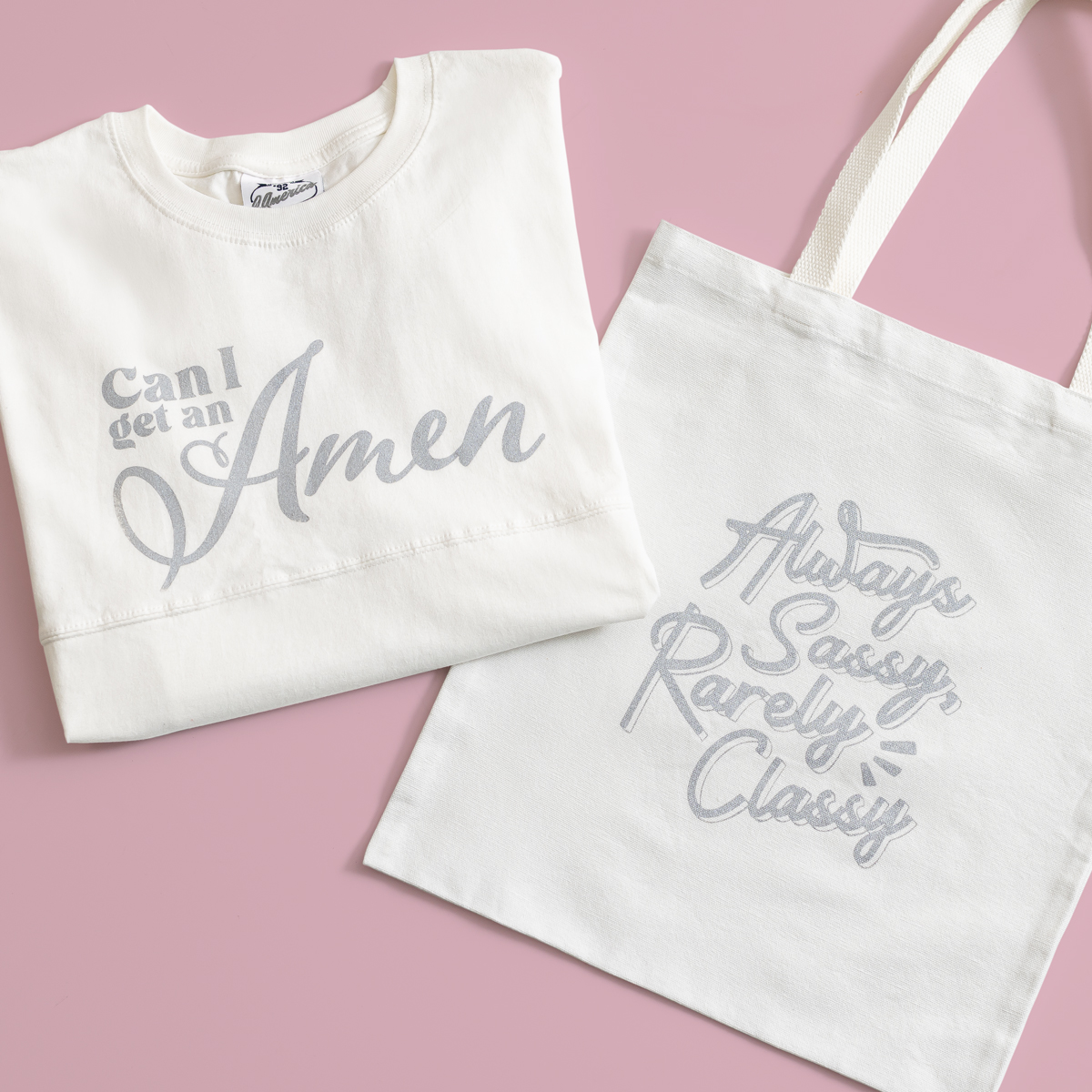 A white sweatshirt reading "Can I get an Amen" is folded next to a white tote bag that says "Always Sassy, Rarely Classy," both displayed on a pink background.