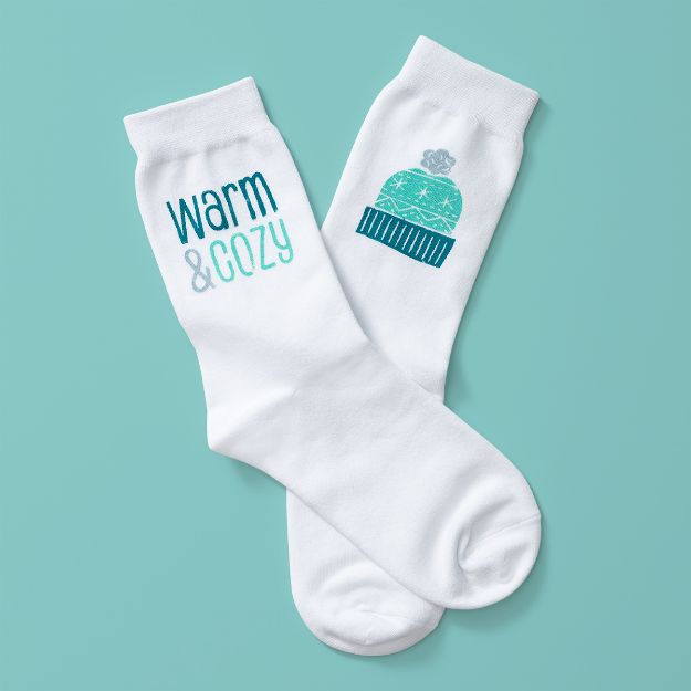 Two white socks on a teal background; one sock features the words "warm & cozy" in blue and teal, and the other has a blue and teal knit hat graphic.