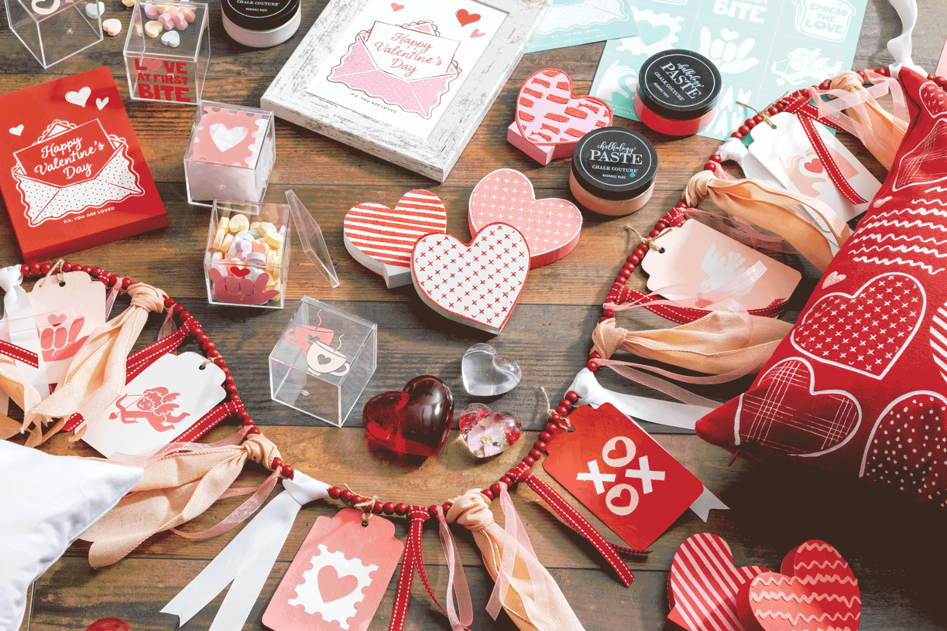 A variety of Valentine’s Day-themed items, including heart-shaped decorations, gift tags, candies, small jars, a garland, and cards, all in red, pink, and white, arranged on a rustic wooden table.