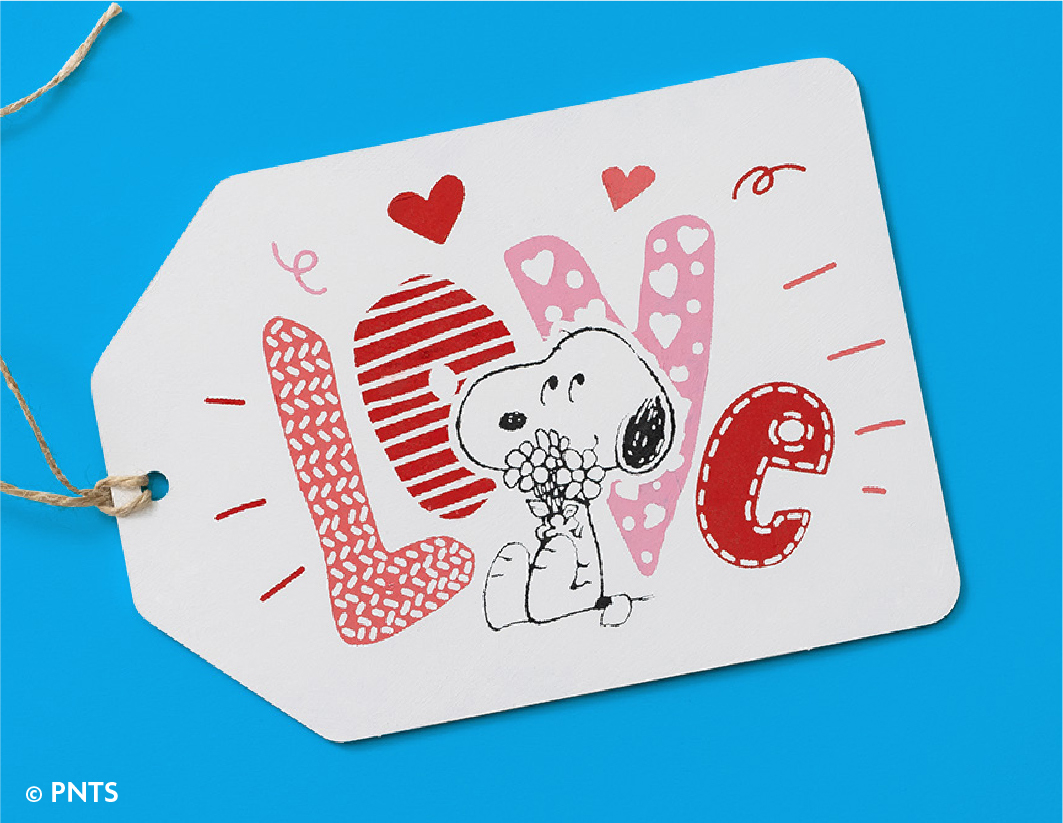 A gift tag with the word "LOVE" in red and pink patterns, featuring Snoopy holding flowers. Two red hearts and decorative lines surround the illustration. The background is blue.