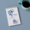 A spiral notepad with a vintage blue botanical illustration and the text "Dated 1849" lies on a blue surface next to a turquoise mug filled with black coffee.