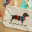 A canvas tote bag with a print of a dachshund wearing a colorful, textured sweater and a red collar, with paw prints below. Embroidery thread, a stencil, and crafting supplies are nearby on the wooden surface.