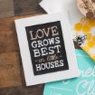 A framed sign with the phrase "Love grows best in little houses" written in white and pink letters, placed on a wooden surface next to a bee-themed towel and a teal sign.