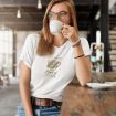 A woman with glasses sips from a white mug in a modern cafe. She is wearing a white t-shirt that says, "just a girl that loves coffee" with a graphic of a coffee cup. She looks relaxed and thoughtful.