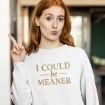 A woman with long red hair wearing a white sweatshirt that says "I COULD be MEANER" makes a duck face and holds up a peace sign with her right hand. She stands indoors with a blurred background.