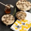 Wooden boards with engraved honeycomb and bee designs are arranged on a dark surface next to a jar of honey with a dipper, a sprig of rosemary, and a white and yellow cloth with bee patterns.