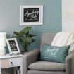 A cozy living room corner with a gray armchair, green "Homebody Club" pillow, plant, side table, lamp, framed sign on the wall, and another framed quote on the table, against a blue accent wall.