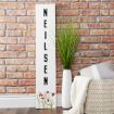A white wooden sign with the name "NEILSEN" written vertically in black letters stands against a brick wall, next to a tall white vase with grass and a cream-colored knit blanket draped over a woven chair.