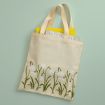 A beige canvas tote bag with light handles features a design of green stems and white flowers. The bag contains a yellow item and a partially visible book or magazine. The background is a light green color.