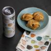 A blue plate with three pastries sits on a white napkin decorated with colorful cup illustrations, next to a travel tumbler featuring a similar cup pattern, all on a dark surface.