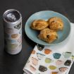 A blue plate with three pastries sits on a white napkin decorated with colorful cup illustrations, next to a travel tumbler featuring a similar cup pattern, all on a dark surface.