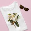 A white t-shirt with a large botanical illustration of a magnolia flower is neatly folded on a pink background next to a pair of dark sunglasses.