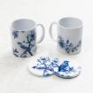 Two white mugs with blue floral and bird designs, placed on a light surface. In front of the mugs are two round coasters with matching blue bird and flower illustrations.