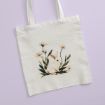 A white tote bag with an illustration of a white rabbit sitting among green leaves and white daisies, placed on a light lavender background.