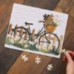 A nearly completed jigsaw puzzle depicts a bicycle with a basket of flowers next to a wooden fence. One piece is missing, and a hand is holding a puzzle piece above the puzzle. Three other pieces lie on the table.