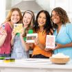 Four smiling women stand together in a bright kitchen, each holding a decorative sign with positive messages. Popcorn and small paint containers are on the counter in front of them.
