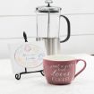 A pink coffee mug with the words "just a girl that loves coffee," a French press, and a small sign on a stand that says "All are welcome at our table," sit on a white countertop.