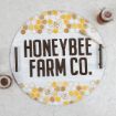 A round tray with handles features honeycomb patterns, bee illustrations, and the bold text "HONEYBEE FARM CO." Two jars of honey and a honey dipper are placed next to the tray on a light-colored surface.